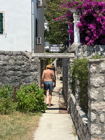 A shirtless man wearing a straw hat and blue shorts walks along a paved path next to a stone wall. He is facing away from the camera, with his right hand resting on his hip and his left arm slightly bent. The path appears to be in an outdoor setting, possibly a coastal town given the presence of lush green foliage and flowering plants. To the left of the path, a white building with a dark green window shutter is visible. A sign with black lettering on a white background reads "ULICA MIRISA MIMOZE". The time of day appears to be sunny, with distinct shadows cast on the ground. The overall scene evokes a relaxed, summery atmosphere.