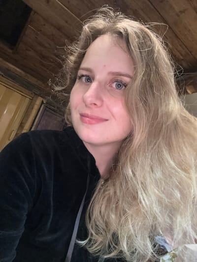 The image is a close-up, indoor selfie of a young Caucasian woman with blonde, wavy hair and blue eyes. She is wearing a black hooded jacket. The background consists of wooden paneling on the ceiling and walls, suggesting a rustic or cabin-like environment. The lighting is warm and appears to be artificial. There are no other people or significant objects visible in the frame. The image does not provide any specific location details within Severnyy, Russia.