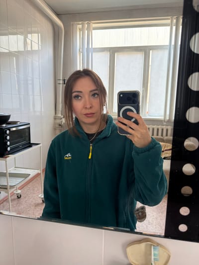 A young woman with shoulder-length brown hair is taking a selfie in a bathroom mirror. She is wearing a dark green fleece jacket with a yellow zipper pull and a silver chain necklace. Her eyes are blue-green, and she is wearing makeup. In the background, reflected in the mirror, is a window with vertical blinds, a radiator, and a couch with striped cushions. To the left, also reflected, is a small black electric oven on a white rolling cart. The bathroom walls are tiled with white square tiles. The floor appears to be linoleum with a pinkish-tan pattern. A black frame with circular cutouts is visible to the right of the mirror. The scene is likely in a residential bathroom.