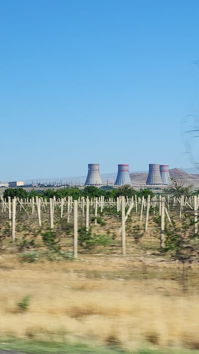 In Arshaluys, Armenia, four large cooling towers stand prominently against a clear blue sky, characteristic of a power plant. In the foreground, a vineyard with rows of wooden posts and wires stretches across the landscape. Beyond the vineyard, a line of green trees separates the agricultural area from the industrial structures. The ground in the immediate foreground is covered in dry, brown vegetation, suggesting a warm climate. The overall scene depicts a juxtaposition of agricultural land and industrial infrastructure in a rural setting.