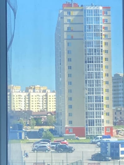 This image captures a residential scene in Nefteyugansk, Russia, on a clear, sunny day, as viewed through a window with a blue curtain on the left. In the foreground, a paved parking lot is visible with several cars, including a gray sedan, a dark SUV, and a white van. Two individuals, appearing to be a woman and child with a stroller, are walking across the parking lot.
Dominating the midground and background are multi-story buildings. A tall, modern apartment building with a light beige facade and prominent glass-enclosed balconies stands on the right, adorned with yellow and red accents near its rooftop. To the left of this building, a row of lower apartment buildings, also with light beige exteriors and yellow accents, stretches across the landscape. In the far background, another tall building with a grayish facade is visible. The overall environment suggests a modern urban or suburban residential area.