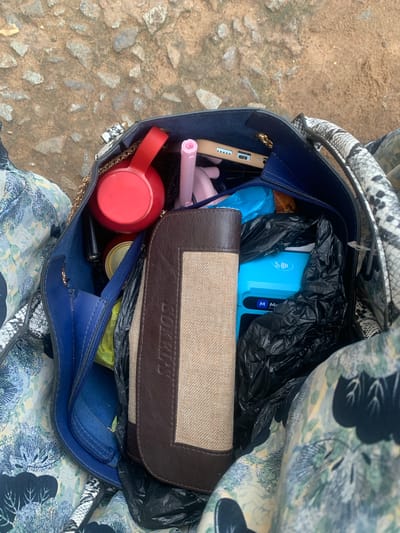 A high-angle, full shot captures the interior of a dark blue tote bag with a snakeskin patterned handle, resting on a textured, rocky ground. The bag is filled with various items, including a red circular object, a pink makeup item, a light brown wallet with a textured beige insert, a blue card reader, and a can. The ground appears to be dirt and gravel, consistent with an outdoor urban environment. The overall scene suggests a snapshot of everyday items within a personal bag in Elelenwo, Nigeria.