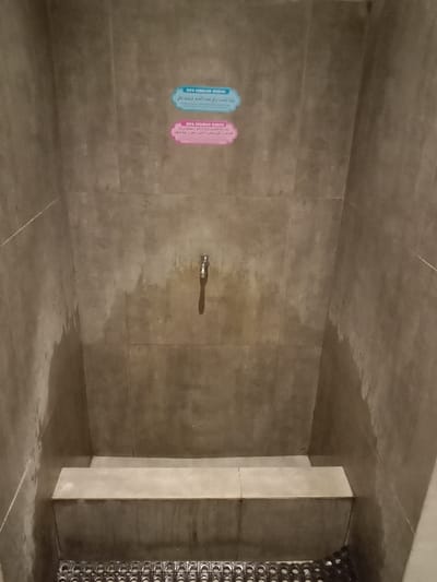 The image shows a small, grey-tiled washroom with a single faucet on the wall. There are two signs, one blue and one pink, with Arabic text and the Indonesian phrase "DOA SEBELUM WUDHU" and "DOA SESUDAH WUDHU" on them. The wall is wet from water dripping from the faucet. There is a bench beneath the faucet and a dark grey mat on the floor. No people are present in the image. The location is within the city of Kecamatan Ngaliyan, Indonesia, which is a district of Semarang, with latitude -7.013 and longitude 110.338.