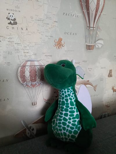 The image shows a stuffed green dragon toy with white spots on its belly, positioned in front of a wall mural of a world map. The map features illustrations of hot air balloons, an octopus, a panda, a dolphin, and various animals like a bear and a deer. The dragon has wings and is slightly leaning to the right. In the lower left corner, a wooden toy airplane is visible. The background is a textured wallpaper with the map. The scene is indoors, on what appears to be a sofa or upholstered surface. There is no indication of the city of Biaroza, Belarus in the image.