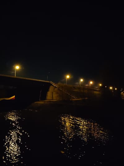 The image depicts a nighttime scene in Podosinki, Russia, featuring a bridge illuminated by streetlights. The bridge spans a body of water, with light reflecting off its surface. The surrounding area appears dark, with the bridge and its lights serving as the primary focus.