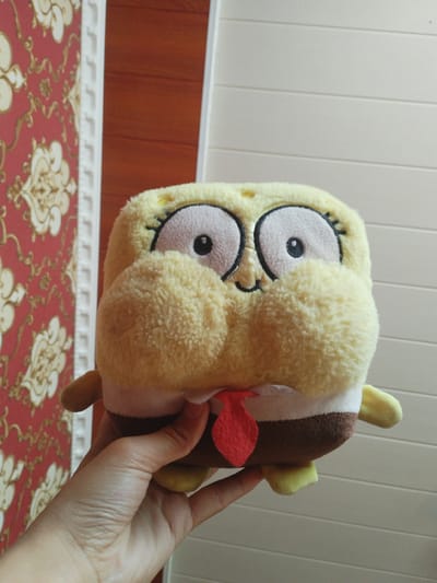 A hand holds a plush toy resembling SpongeBob SquarePants. The toy is yellow and has oversized, white-rimmed eyes with black pupils, rosy cheeks, and a red tie. It is being held in front of a wall with red wallpaper featuring a gold floral pattern and a white decorative border. To the right of the wallpaper is a light-colored wall with horizontal paneling. The lighting suggests an indoor setting during the daytime.