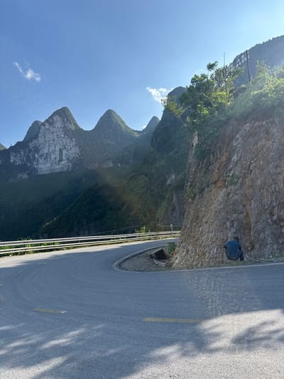 A road cuts through a mountainous landscape in Đồng Văn Commune, Vietnam. The road, marked with a yellow line, curves along the edge of a steep rock face. A metal barrier runs along the edge of the road, separating it from the drop-off. Further up the mountain are dense forests and rocky peaks under a clear blue sky. A dark figure is visible perched on the side of the rock face, close to the road.