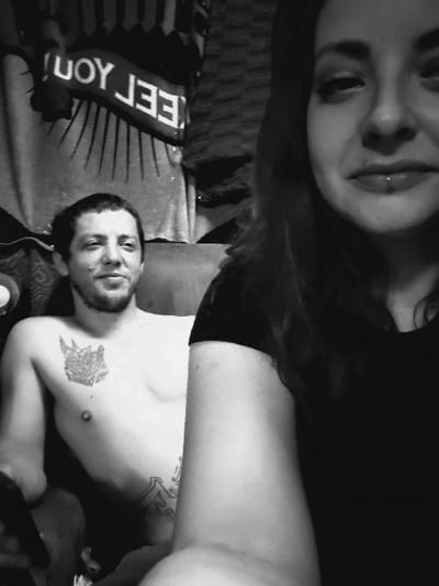 The image shows a man and a woman in an indoor setting. The man, who is shirtless and has tattoos on his chest and arm, is seated in a chair. He is holding a controller in his left hand. The woman is standing behind him, wearing a black shirt. Her right arm is partially visible. Her face is showing, with a slight smile and a piercing in her lower lip. The image appears to be taken in a living room or bedroom, as there is a couch in the background. The wall behind the man has a piece of cloth with the inscription "FEEL YOU" on it. The image was taken in North Highlands, California, United States. The location, based on the coordinates provided, is a residential neighborhood. The overall ambiance of the image is relaxed and casual.