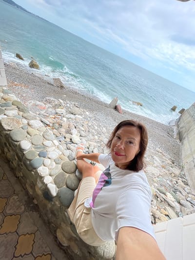 A woman with shoulder-length brown hair and dark eyes is taking a selfie on a pebbly beach in Abaata, Abkhazia. She is wearing a white t-shirt with a colorful abstract design on the front and light-colored shorts. Her left leg is bent with her foot resting on a low stone wall made of rounded grey and white stones. The beach is covered in small grey and white pebbles, with some larger rocks and concrete blocks scattered along the shoreline. The turquoise water of the sea is visible in the background, with waves breaking on the shore under a cloudy sky. To the left of the woman, a patterned tiled surface is visible.