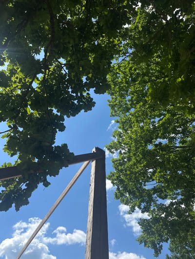 The image depicts an upward view of a wooden structure against a bright blue sky with scattered white clouds. Lush green foliage from trees, likely oak based on the leaf shape, frames the top and sides of the frame. The wooden structure appears to be part of a pergola or similar outdoor fixture, with thick, weathered beams forming an angled frame. One large, dark wooden post stands prominently in the lower central portion of the image. The scene is set outdoors, suggesting a park, garden, or rural area within Завалочыцкі сельскі Савет, Belarus. There are no people or animals visible in the image.