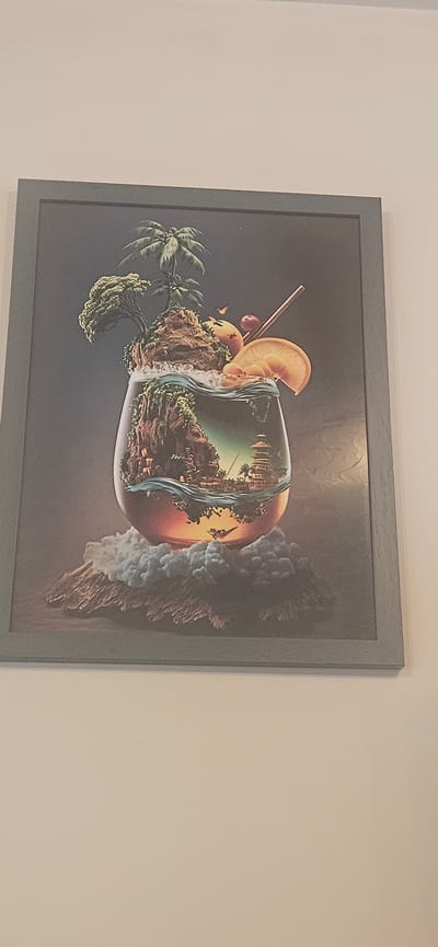 A framed painting hangs on a light beige wall. The painting depicts an surreal, tropical-themed image. It shows a cocktail glass containing a landscape featuring palm trees, cliffs, and a waterfall. A slice of orange and a cherry garnish the glass. A straw extends from the glass. On the right side of the glass, a cloud formation is present. The painting is enclosed in a gray frame.