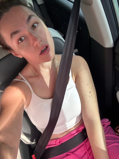 An individual is seated in the passenger seat of a vehicle, wearing a white tank top and pink shorts. A black seatbelt is fastened across their chest. The individual's face is visible, with their eyes wide open and mouth slightly agape. Sunlight is casting shadows on their face and arm. The interior of the car is dark, with a light-colored pillar visible behind the individual. The image appears to have been taken from a slightly high angle, looking down at the individual.
