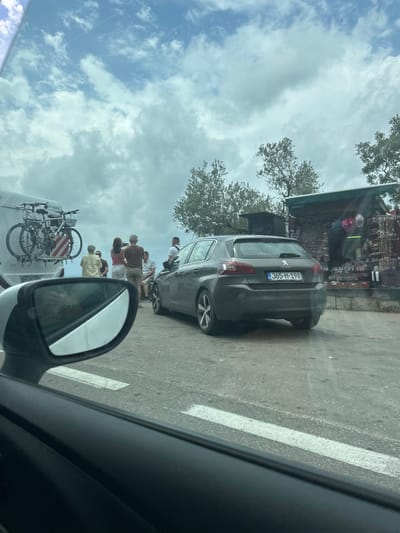 The image, taken from within a vehicle, depicts a casual daytime scene along a roadside in Pržno, Montenegro. The sky above is bright and mostly cloudy.
In the mid-ground, a dark grey Peugeot hatchback is parked on the right side of the road, facing away from the camera. Its Montenegrin license plate, "J65-M-198," with "MNE" visible on a blue strip, is clearly legible. Behind the car, a small, rustic roadside stall or shop is visible, displaying various goods, possibly souvenirs or handicrafts. Trees with sparse foliage frame the upper and middle sections of the scene.
Several people are gathered on the roadside. Some individuals are standing near the parked Peugeot, while others are closer to the left, partially obscured by the viewer's car's side mirror. Through this mirror and in the lower-left corner, part of a white van is visible, equipped with a bicycle rack holding two bicycles. The people appear to be casually dressed, suggesting they might be tourists or locals making a stop.
The foreground shows the dark frame of the viewer's car window and its side mirror. The road surface features distinct white lines marking the edge. The overall impression is a relaxed, perhaps scenic, stop along a route, likely for browsing the roadside stall or enjoying the location.