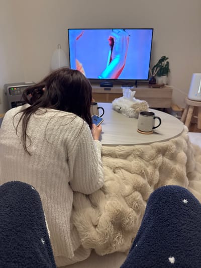 The image portrays an indoor domestic scene within Utsunomiya, Japan. In the immediate foreground, the extended legs of an unseen individual are visible, clad in dark blue, fuzzy socks patterned with small white polka dots. A second person, with long, dark brown hair, is seated on the floor with their back to the camera, wearing a white, textured, long-sleeved top. This individual is actively operating a light-colored smartphone held in both hands, while also facing a large, flat-screen television positioned on a low, light-colored wooden media cabinet against a white wall.

The television screen displays an abstract or artistic image featuring vibrant pink, blue, and purple hues, depicting what appears to be a stylized human torso or limb with high contrast. Directly in front of the seated person is a kotatsu, a low, round table with a light-colored wooden top. A thick, beige, faux-fur blanket drapes over the table, characteristic of a kotatsu. On the tabletop, a white, fluffy, animal-shaped tissue box cover is positioned between two mugs: one dark-colored with a silver rim, and another light-colored with a dark handle and silver rim, resting on a wooden coaster.

In the background, to the left of the television, a grey, vertical appliance (possibly a heater) and a white, translucent, inverted U-shaped lamp are visible. To the right of the television, a small green plant in a white pot rests on a wooden stand, alongside a white, modern humidifier placed on a small wooden stool. The overall environment suggests a cozy, residential setting with soft ambient lighting.