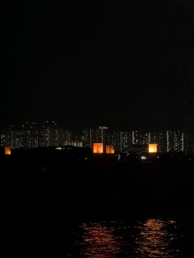 This is a nighttime, horizontal, eye-level, medium shot of a reflective skyscraper in Nanshan District, China. The building is tall and slender, with many windows, some of which are lit with an orange glow. The building's reflection can be seen in the dark water below. The water is disturbed, with streaks of light that appear to be reflections of distant lights or possibly fireworks. The overall mood is mysterious and serene. The image is well-lit, with the skyscraper being the main subject, and the reflections in the water adding a sense of depth and dimension. The time of day is night, and the weather is clear. The location is a city, likely near a body of water. No people or text are visible in the image.