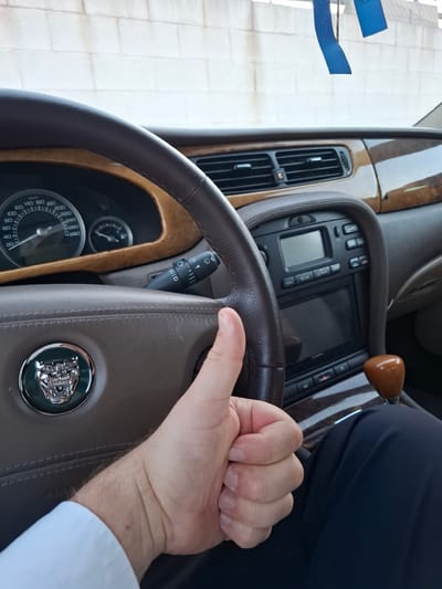 Here is a forensic description of the image:
The image shows the interior of a vehicle, specifically the steering wheel of a Jaguar. The steering wheel is brown leather with a central emblem. The dashboard is visible in the background, featuring a wood trim, air vents, and a display screen. The gauge cluster can also be seen, showing the speedometer, marked in km/h. A hand is resting on the steering wheel, wearing a white shirt. The background also shows a glimpse of the exterior with white tiled pavement. The image does not include enough information to determine if the image was taken in Quart de Poblet, Spain.
