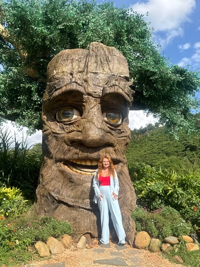 A young woman with long, wavy red hair stands in front of a large, carved tree trunk that resembles a face. The tree has large, expressive eyes and a wide, toothy grin. The woman is wearing a red tank top and light blue loose-fitting pants and jacket. She is standing on a path made of stone and gravel, with lush green foliage and small flowers surrounding the base of the tree. In the background, a hillside covered in dense green vegetation slopes upwards under a bright blue sky with scattered clouds. This scene is located in Thôn 1, Vietnam, likely within a garden or botanical attraction.