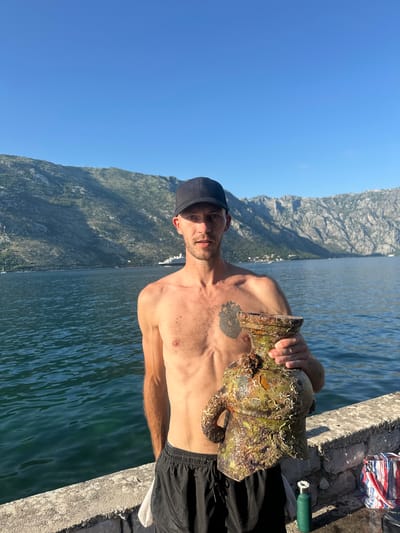 An image taken outdoors in Prčanj, Montenegro, depicts a male figure standing beside a body of water with a mountainous background under a clear blue sky.
The central figure is an adult male, appearing to be in his late 20s to early 30s, with fair skin and short facial stubble. He wears a black baseball cap and black athletic shorts with a visible grey waistband and white drawstring; he is shirtless. A tattoo resembling an anatomical heart with root-like elements is visible on his left pectoral. He holds a large, irregularly shaped object, heavily encrusted with marine growth, predominantly dark green, brown, and off-white. The object appears to be a relic, possibly a vase or urn, with a wide body, a narrower neck, and two arm-like protrusions, all covered in barnacles and other marine organisms. His gaze is directed forward, towards the camera, with a neutral expression.
The man stands on a light-grey concrete or stone seawall, constructed from stacked blocks. To his right, on the wall, a striped bag (red, white, and blue) and a small, dark green cylindrical bottle or container are present. A white cloth or towel is partially visible, tucked into his shorts on his right side.
The background features a large, calm body of dark blue-green water, indicative of a bay or fjord. Steep, rocky mountains, partially covered in sparse vegetation, rise directly from the water on both the left and right sides of the frame. In the distance, a large white cruise ship or yacht is visible on the water towards the center-right, and a smaller sailboat is present further left. The sky is uniformly clear and bright blue, suggesting fair weather and strong sunlight, casting visible shadows on the man's face and the object he holds.