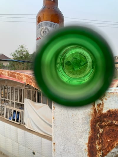 The image depicts a view through the neck of a green bottle, possibly a beer bottle, capturing an outdoor scene. A portion of a brown bottle is visible on the left. The interior view through the green bottle neck shows greenery, possibly foliage. The surrounding environment includes a white tiled surface, a white covering, metal structures, and an aged metal surface with rust. The sky is visible in the distance, suggesting an outdoor setting.