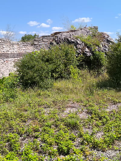 The image shows the remains of an ancient Roman fortress in Garmen, Bulgaria. The fortress is made of stone and is partially overgrown with vegetation. The sky is blue with some clouds. The fortress is located on a hill overlooking the surrounding landscape.
