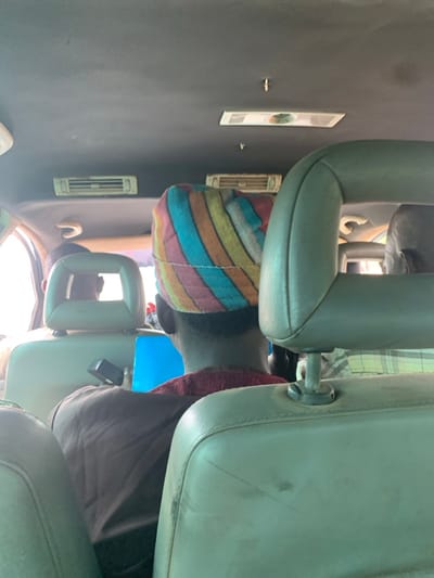 The scene depicts the interior of a vehicle, likely a passenger van or bus, in Alizaga, Nigeria, viewed from a rear seat looking forward. Three individuals are visible from behind. In the immediate midground, a person wears a distinctive multi-colored striped head covering in shades of pink, orange, yellow, green, and blue, over a dark reddish-brown garment. To their left, another individual wears a dark garment, holding a bright blue object, possibly a folder or tablet, and is seated in front of a light-colored headrest with a rectangular cutout. To the right of the central figure, a third person's head and shoulder are partially visible, wearing a light-colored plaid shirt. The vehicle's interior features a dark grey ceiling with overhead air conditioning vents, including two elongated rectangular vents and a central circular vent panel. The seats are light-colored, possibly beige or light green, showing signs of wear and minor discoloration. Natural light enters from the left side, indicating daylight outside.