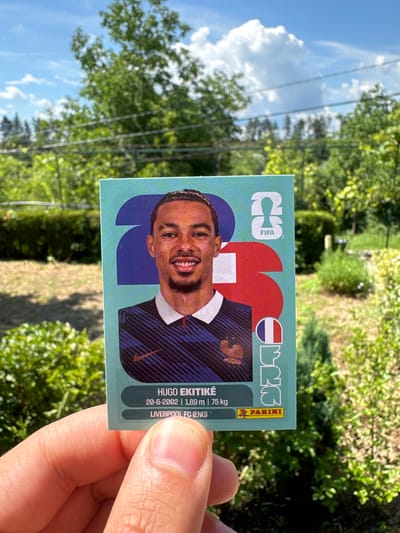 An extreme close-up outdoor shot shows a hand holding a soccer trading card featuring Hugo Ekitiké, a French footballer. The card displays his image, name, birthdate (20-6-2002), height (1.89 m), weight (75 kg), and club (Liverpool FC (ENG)). The background is blurred, but suggests a natural environment with trees, grass, and a bright sky with clouds. The location is identified as Prisovo, Bulgaria.