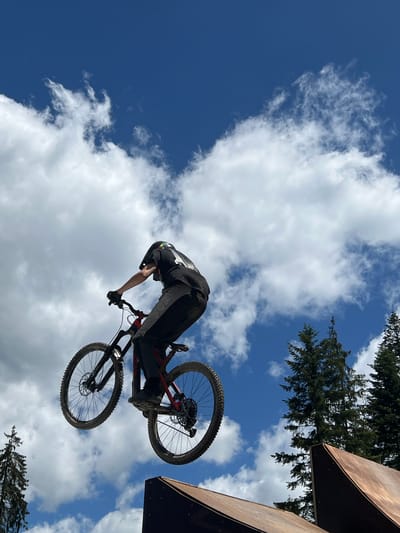 A person wearing a helmet, black t-shirt, and grey pants with black accents is riding a mountain bike in mid-air. The bike is red and black. The person is leaning forward, with their arms extended to the handlebars. The background is a bright blue sky with white, fluffy clouds. Trees are visible in the upper right and lower left corners of the image, and a wooden ramp structure is in the lower right corner. The image appears to have been taken at a bike park, possibly in the area surrounding Samokov, Bulgaria, given the presence of trees and hilly terrain typical of such locations.