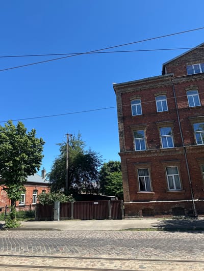 The image captures a street scene in Liepaja, Latvia, on a clear, sunny day. In the foreground, cobblestone pavement is visible, with what appear to be tram tracks running horizontally across the bottom. A brown wooden fence lines the sidewalk, separating it from a property with a brick building and trees. To the right, a multi-story brick building with numerous windows dominates the frame, its facade exhibiting traditional architecture. Overhead, power lines crisscross the bright blue sky. The overall impression is that of a residential street in an older European city.