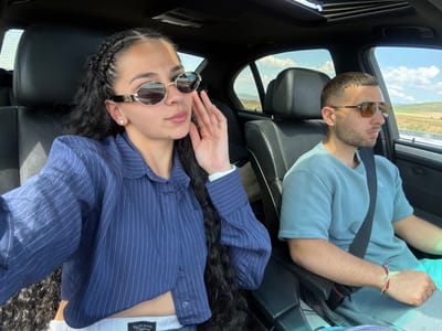 Two individuals are seated in the front seats of a car, traveling on a highway. The female passenger on the left is wearing a navy blue pinstripe shirt and sunglasses. Her hair is styled in braids. The male driver on the right is wearing a light blue textured t-shirt and sunglasses. The car's interior, including leather seats and a sunroof, is visible. The exterior view from the car windows shows a landscape with fields and a cloudy sky, consistent with the rural environment of Polsko Padarevo, Bulgaria.