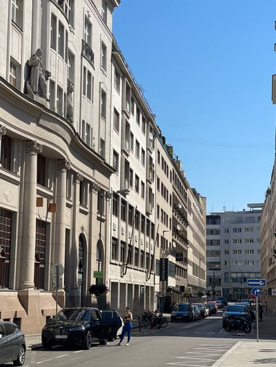 The image shows a street scene in Bratislava, Slovakia, under a clear blue sky. On the left, a grand building with classical architectural elements, including large columns and decorative statues, lines the street. The building is a light color, likely stone or stucco, and has numerous windows. To the right, a series of buildings of varying heights and architectural styles form the other side of the street. These buildings are also light-colored and feature many windows.

In the foreground, a black BMW SUV is parked on the street with its driver's side door open. A woman in blue leggings and a dark top stands next to the open door, appearing to be interacting with the vehicle. Another car, a gray hatchback, is partially visible on the far left. The street itself is paved, with white lane markings.

Further down the street, several other cars are parked and in motion. Traffic signs are visible, including directional signs and parking indicators. Signs for businesses are also present on the buildings. The overall impression is of an urban environment on a sunny day.