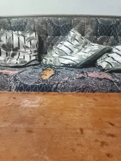 An indoor scene in Cimenyan, Indonesia, displays a sofa or daybed positioned against a plain, light-colored wall. The sofa features a dark, patterned upholstered backrest. Its seat is covered by a dark blue and black patterned blanket or throw with subtle red or pink accents. A significant, irregular tear in this blanket, approximately 15-20 cm in its longest dimension, exposes a lighter brown material underneath. Two pillows with black and white leaf patterns are arranged on the sofa; one on the left features vertical stripes, and the one on the right is partially obscured by a folded, dark green or grey fabric item. The lower half of the image reveals a light brown wooden surface, possibly a table or the front frame of the furniture, exhibiting wear and faint marks. No individuals are present, and no actions are in progress.