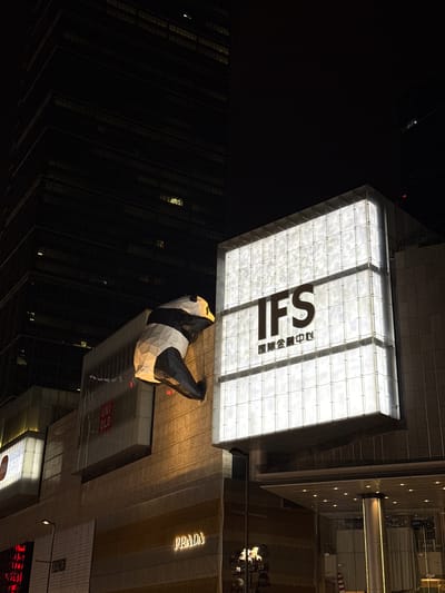 An eye-level, nighttime shot shows the International Finance Square (IFS) building in Jinjiang District, Chengdu, China. A large panda sculpture sits on the exterior of the building, its black and white fur contrasting against the city lights. Adjacent to the IFS signage, luxury brand storefronts like Prada, Uniqlo, and Fendi are visible. The lighting is bright, illuminating the architecture and drawing attention to the storefronts. The darkness of the night forms a stark backdrop to the illuminated scene.