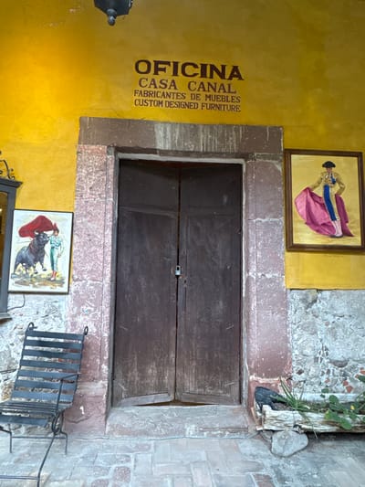 The image shows a building facade in San Miguel de Allende, Mexico, with a prominent wooden door framed by stonework. Above the door is a painting of a bullfighter, while another painting depicting a bullfight is below the door. The building's yellow wall is painted with the words "OFICINA CASA CANAL FABRICANTES DE MUEBLES CUSTOM DESIGNED FURNITURE". A metal chair stands near the door, adding to the scene. The overall architecture reflects the city's colonial style.