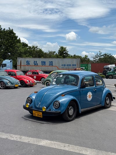 A fleet of vintage Volkswagen Beetles are parked in an outdoor lot, with shipping containers and a blue truck in the background. The cars are various colors, including blue, red, and green. The blue Beetle in the foreground has a yellow license plate reading "22 0846". The scene is illuminated by daylight, with a cloudy sky above. The location is Lao Bảo Commune, Vietnam.