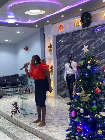 The image depicts an indoor event, likely a Christmas celebration, occurring within a modern hall or church in Gbawe, Ghana.

In the foreground, a dark-complexioned female subject stands barefoot on a light-colored, raised platform with a marble-patterned floor. She wears a vibrant red blouse with ruffled sleeves and a black knee-length skirt, holding a red microphone near her mouth, indicating singing or speaking. To her left, a clear, curved glass podium is visible. A small snowman figurine sits on the lower floor level to the far left.

To the right of the primary subject, an artificial green Christmas tree stands, heavily decorated with multi-colored ornaments (red, green, silver, gold, pink), illuminated with blue string lights, and topped with a white star. Behind the Christmas tree, a dark-complexioned male subject stands, wearing a white collared shirt, dark trousers, and a dark belt. He holds a dark microphone in his right hand. Dark shoes are visible on the floor near his feet. A second female subject, wearing red, is partially visible behind the primary female subject.

The background wall features a dark, possibly marble-patterned section with multiple illuminated white niches displaying vases with red flowers and other decorative items. Above these, a gold "NOEL" sign and a circular Christmas wreath with red accents are mounted. A red heart-shaped decoration is visible on a white wall section to the far left. The ceiling is multi-tiered with recessed white lighting and purple LED strip lighting along the edges. Rows of empty dark chairs are visible in the background to the far left.