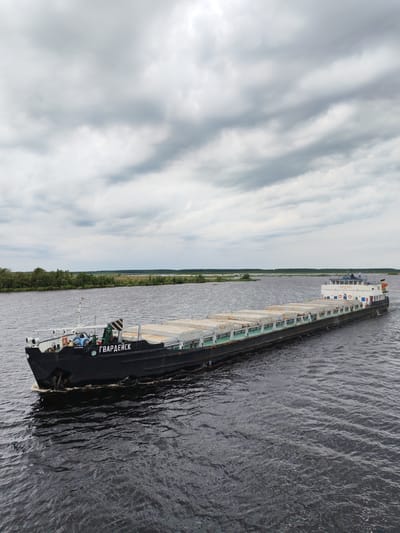 A barge named "ГВАРДЕЙСК" sails on a dark, choppy river under a cloudy sky. The barge, which appears to be a cargo barge, is laden with rectangular, pale-colored containers. The barge is black with a white superstructure at the stern, which features windows and life preservers. A crane is visible on the deck. The riverbanks in the background are lined with green trees. The location is identified as Novokemskii, Russia.