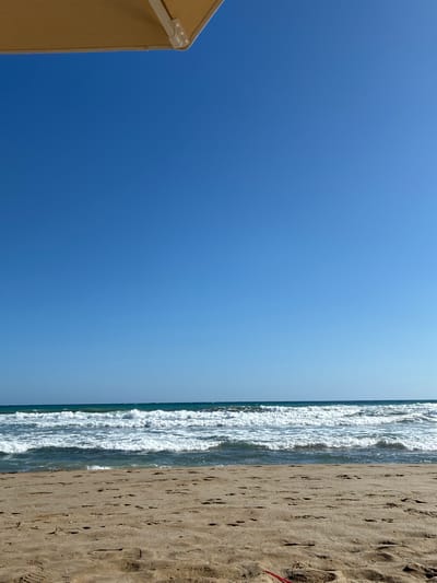 A sandy beach on a clear, sunny day with a bright blue sky and calm ocean. The sand is light brown with visible footprints. The water has gentle waves breaking on the shore, appearing turquoise and clear. In the upper left corner, a portion of a light brown umbrella is visible, providing shade. The scene is tranquil and appears to be in Rethymno, Greece, a popular coastal city. There are no people or animals visible in the image.