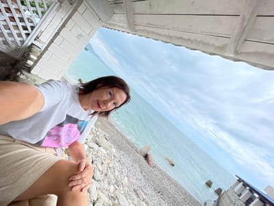 A woman is taking a selfie on a pebble beach in Abaata, Abkhazia. She is wearing a white t-shirt with a graphic print and tan shorts. She is sitting down, with her left hand resting on her left knee. The background shows the sea and a cloudy sky. There are some concrete blocks in the water and on the beach. Part of a white structure with a lattice design is visible on the left side of the image.