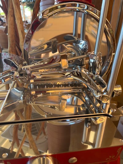 The image shows a close-up of a polished chrome-plated meat slicer, likely in a commercial setting. The slicer has a large circular blade, a carriage mechanism, and several levers and handles, all reflecting the surrounding environment. The background is blurred, but appears to be an indoor space with natural wood elements and possibly tables and chairs, suggesting a restaurant or deli. There are no people visible in the immediate foreground. The image's context suggests it was taken in Kovačko Polje, Montenegro, though the specific location within the city is not discernable from the image itself.