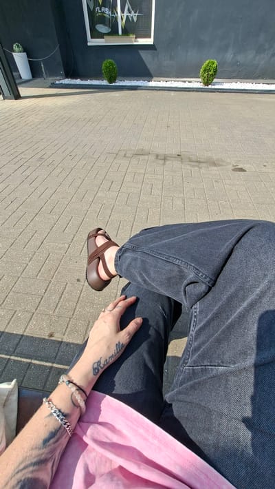The image shows a person sitting outdoors, with their legs crossed, wearing dark wash jeans and brown sandals. The person's left arm is visible, adorned with several bracelets and a tattoo on the forearm. The background consists of a paved area with a dark wall, a window displaying a "FLASH ULTRA ENERGY" logo, two small green bushes, and a white decorative rock bed. The scene is lit by sunlight, casting shadows. The location is likely in the city of Smalyavichy, Belarus, given the prompt.
