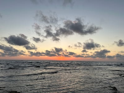 A serene sunset scene unfolds over the Mediterranean Sea near Haifa, Israel. The sky transitions from a pale blue at the top to warm hues of orange and yellow near the horizon, where the sun is setting. Scattered, dark clouds drift across the sky, some tinged with pink from the setting sun. The sea is choppy, with small waves lapping against rocky outcrops and the shore, visible in the foreground. The overall mood is peaceful and tranquil, characteristic of a coastal evening. No people or text are visible in the image.