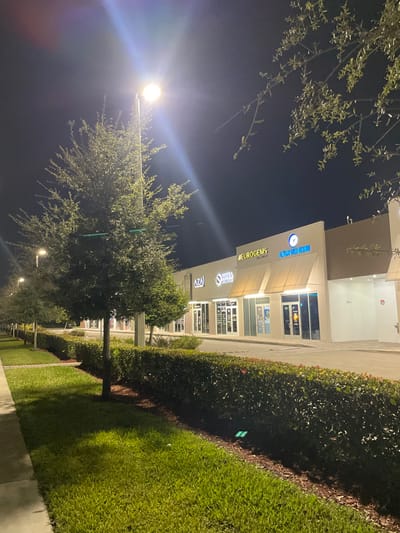 A night shot depicts a building's facade with several business signs, including "Euro Gems" and "Medical Arts Division." Illuminated windows suggest stores or offices inside. Streetlights cast a glow on the scene, highlighting nearby trees. Green hedges and a manicured lawn line the sidewalk, adding a touch of nature to the urban setting. The overall perspective is slightly tilted, creating a dynamic view of the streetscape.