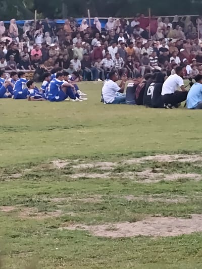 The image depicts an outdoor event, likely a sports match, with a crowd gathered in the background and a group of individuals seated in the foreground on a grassy field. The crowd in the background consists of many people, some standing and some sitting, under a blue and white tarp structure. They are dressed in various casual attire, with some women wearing headscarves. The seated individuals in the foreground appear to be a team, possibly soccer players, as many are wearing matching blue and white uniforms. Some are sitting cross-legged, while others are leaning back or lying down. The environment is a grassy field with some patches of bare soil. The overall scene suggests a community gathering or sporting event taking place in Desa Meunasah Blang, Indonesia.