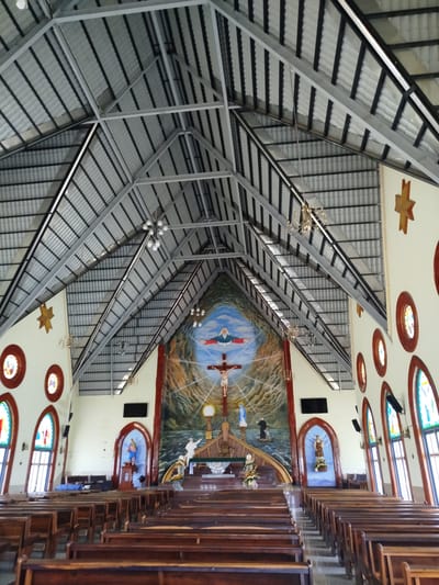 The image shows the interior of a church in K'Rèn, Vietnam. The church features a high, vaulted ceiling supported by exposed metal beams. Pews made of dark wood are arranged in rows facing an altar at the far end. The altar area is adorned with a large mural depicting religious scenes, including a crucifix, figures of saints, and a nautical motif. Stained-glass windows with arched tops are visible on the side walls, casting colorful light into the space. Decorative elements such as stars and chandeliers are also present. The scene appears to be empty of people, suggesting the photograph was taken when the church was not in use.