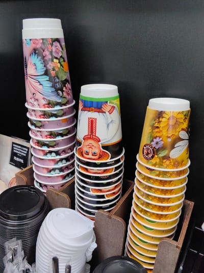 The image shows a close-up view of stacked paper cups in a retail setting, likely a cafe or convenience store. Three stacks of decorative cups are arranged in wooden dispensers. The leftmost stack features cups with a butterfly and floral design. The middle stack displays cups with an illustration of a person in traditional clothing, a smiling face with a straw hat, and a red embroidered pattern. The rightmost stack has cups adorned with sunflowers and a bee. Next to the stacked cups are containers of black and white lids, and some plastic-wrapped cutlery. A label with Cyrillic text and "79.99 РУБ." is visible to the left of the stacks. The background is a plain dark surface. The scene appears to be indoors, with artificial lighting. The location is identified as Myadzyel, Belarus. There are no people visible in the image.