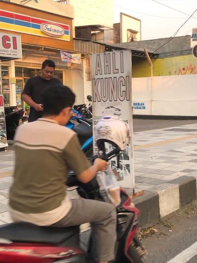 Two men are standing near a scooter on a sidewalk next to a street. One man, wearing a black t-shirt, is looking down at his phone. The other man, wearing a green and white t-shirt, is partially obscured by the scooter. A white helmet is visible on the scooter.

In the background, there is a sign that reads "AHLI KUNCI" (Key Expert) and "BERAS PARKIR" (Rice Parking). To the left, there is an Indomaret convenience store with its distinctive red, blue, and yellow logo. The scene appears to be a street in Sidoarjo, East Java. The lighting suggests it might be late afternoon or early morning. The overall atmosphere is casual, with the individuals engaged in their own activities. The sidewalk has a patterned tile design, and the street is paved with asphalt.
