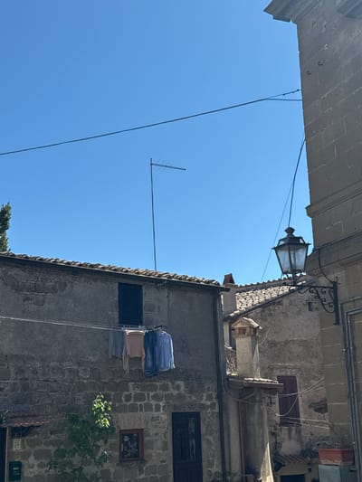 An upward-angled, exterior daytime shot captures a narrow street scene in Castelnuovo di Porto, Italy. On the right, the facade of an old stone building dominates, featuring small, shuttered windows, exposed brick, and a framed picture. Laundry hangs on a rack near the building's side. A portion of the blue sky is visible, with a television antenna extending towards the left. Overhead, the edge of a building is partially visible, obscuring more of the sky. A black, decorative street lamp is mounted on the building on the right. Wires connect the lamp to the building and run across the open space.