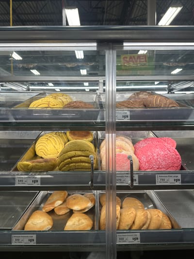 This image depicts the interior of a refrigerated glass display case in the bakery section of a supermarket, likely located in McAllen, United States of America. The case is filled with multiple tiers of various Mexican sweet breads, commonly known as "pan dulce," displayed in metal trays with clear plastic inserts.
From top to bottom, the visible shelves contain:
* **Top shelf (partially visible reflections):** Trays of yellow and brown conchas.
* **Second shelf:** On the left, a tray holds yellow and brown "Concha Large" pastries. On the right, a tray holds brown "Concha Large" pastries. Each is priced at $128.
* **Third shelf (middle):**
* **Left section:** A tray contains numerous yellow "Yellow Concha Large" pastries, characterized by a shell-like scored topping. Some lighter, rounder pastries are also present. A label indicates "540 cal Per 1 Piece" and "$128."
* **Right section:** This section is divided. The left portion holds several light pink, sugar-dusted "Chamuco Pink Large" pastries, labeled "570 cal Per 1 Cookie" and "$128." The right portion holds several vibrant pink, textured "Polvoron Pink Large" pastries, labeled "730 cal Per 1 Cookie" and "$128."
* **Bottom shelf:**
* **Left section:** A tray contains approximately six crescent-shaped "Pineapple Empanada Large" pastries, labeled "510 cal Per 1 Pastry" and "$128."
* **Right section:** A tray contains approximately seven crescent-shaped "Cream Cheese Empanada Large" pastries, labeled "550 cal Per 1 Pastry" and "$128."
The background beyond the display case shows a commercial ceiling with exposed metal beams and overhead fluorescent lighting fixtures. A yellow sign with the red text "SAVE!" is partially visible as a reflection in the upper right glass panel of the display case. No people are visible in the scene.
