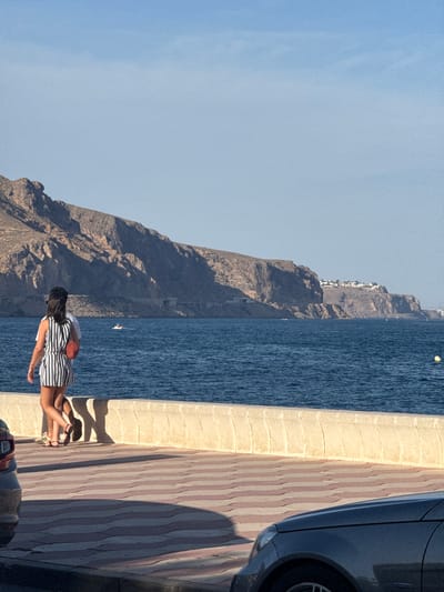 Two individuals, one wearing a striped dress and the other in white, walk along a sunlit promenade. The promenade features a textured, light-colored wall separating it from the deep blue sea. The sea is relatively calm with gentle waves, and a small boat is visible in the distance. Beyond the sea, a rugged, rocky mountain range dominates the horizon, with a few scattered buildings visible near its crest. The promenade itself is paved with interlocking tiles in a pattern of red and cream. A dark-colored car is partially visible in the foreground, casting a shadow on the tiled ground. The sky is clear and light blue, suggesting a warm, sunny day in Aguadulce, Spain.