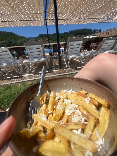 A close-up shot focuses on a bowl of french fries topped with crumbled white cheese, held by a person's hand. A fork is partially visible in the bowl. The background reveals a sunny outdoor scene at a poolside resort, with rows of white lounge chairs under a white mesh canopy. Beyond the chairs, a swimming pool is visible, with lush green hills and trees in the distance under a clear blue sky. This location is likely in Delchevo, North Macedonia, suggested by the context.